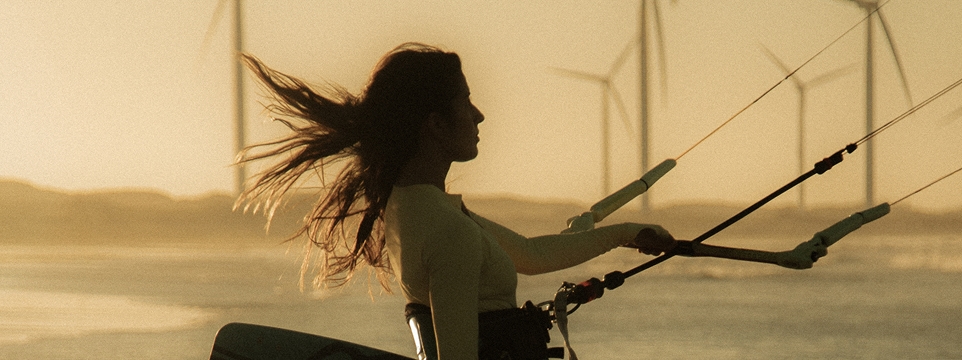 Mulher praticando kitesurf ao pôr do sol, com turbinas eólicas ao fundo.