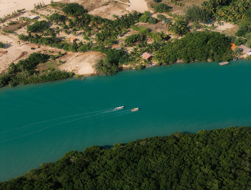 Vista aérea de um rio de águas verde-azuladas com dois barcos, entre vegetação e dunas