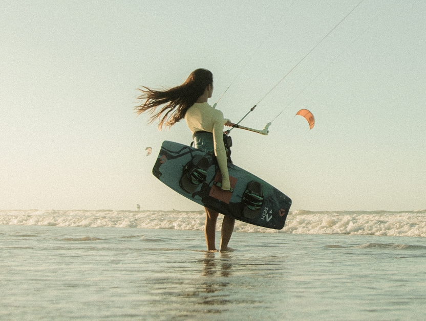 Mulher de costas segurando uma prancha de kitesurf na beira do mar