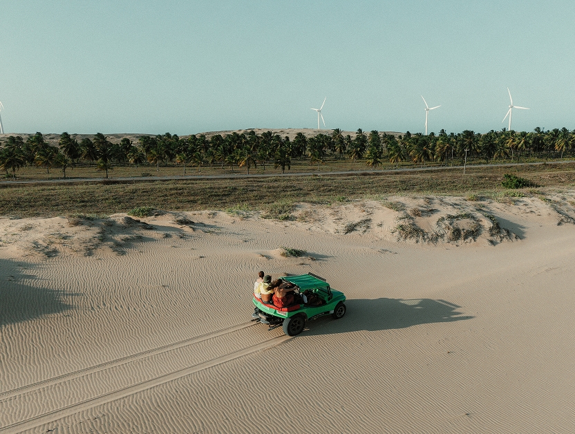 Buggy verde com pessoas cruzando as dunas, com coqueiros e turbinas eólicas ao fundo